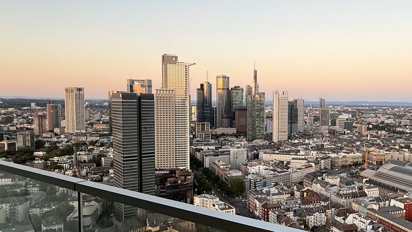 A wide view from the 41st floor balcony across the Frankfurt skyline at dusk, the glass balustrade in the foreground