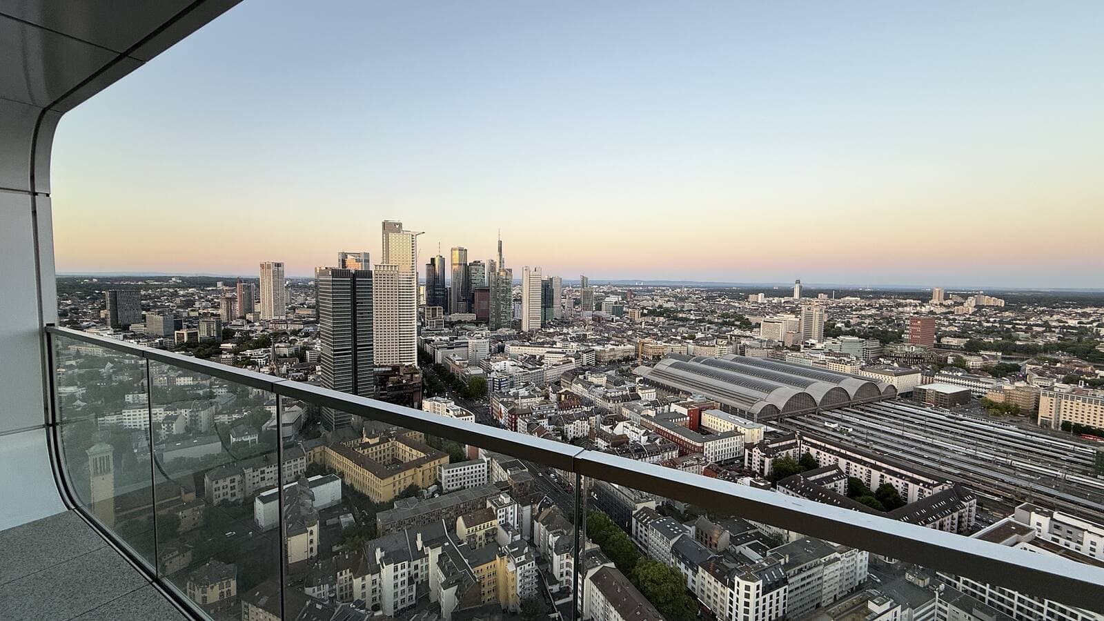 Wrap-around balcony of a Grand Tower residence at dusk, glass balustrade and the Frankfurt skyline beyond